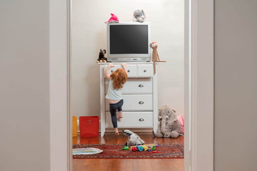 Child hanging on white chest of drawers with TV on top.